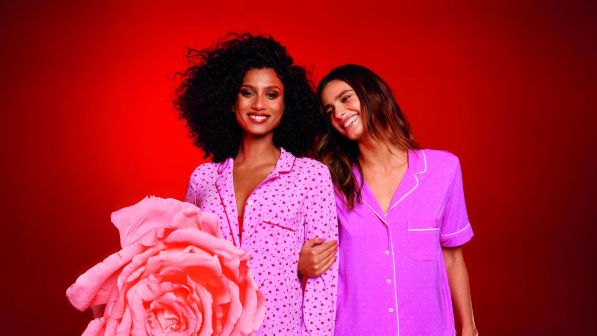 on peut voir sur cette photo en couleurs, deux jeunes femmes habillées en rose qui se tiennent par le bras. il y a une grosse fleur en premier plan et l'arrière plan est rouge.